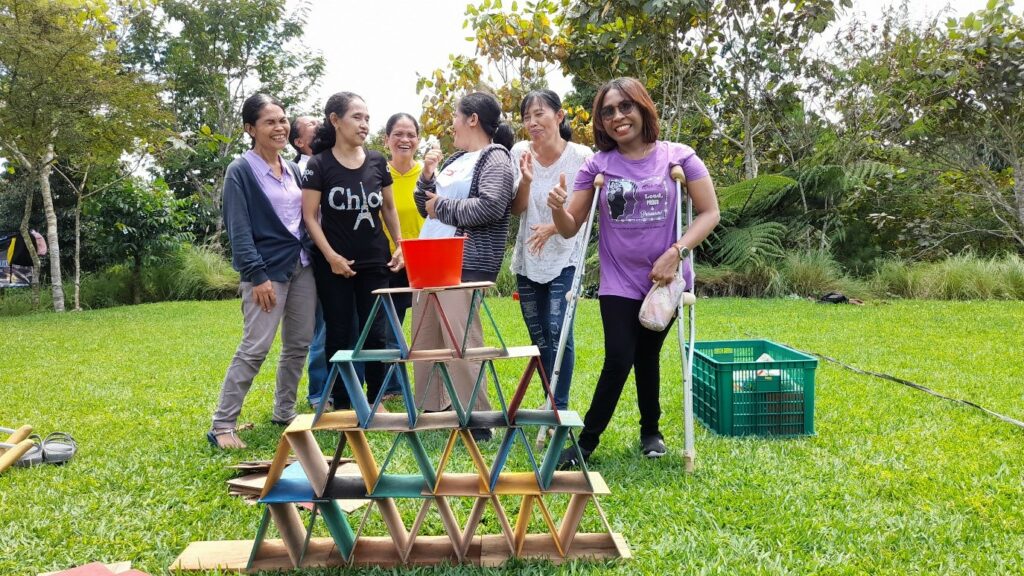 WILD leader Norma uses crutches to stand next to six women participants of WILD-Indonesia at an outdoor park. In front of them on the grass is a tall stack of flat wooden squares arranged in a pyramid shape.