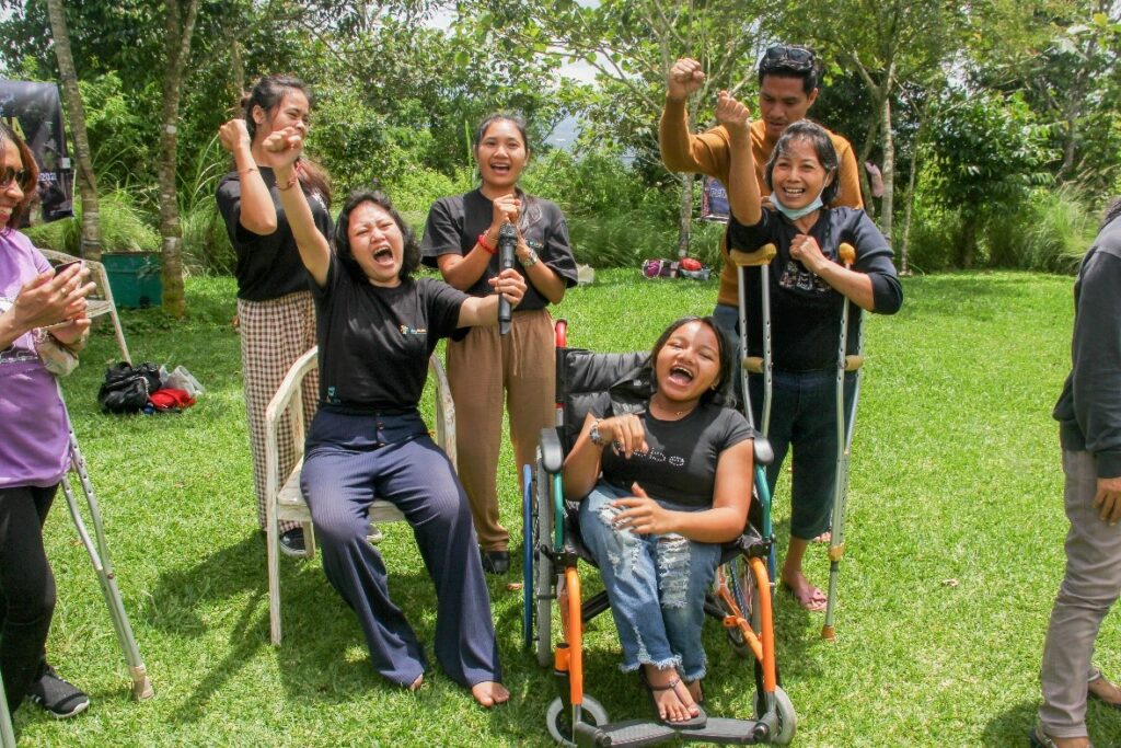 Five women participants of WILD-Indonesia sit and stand together in an outdoor park, holding fists in the air while smiling and shouting. One participants sits in a wheelchair and two participants use crutches to stand.