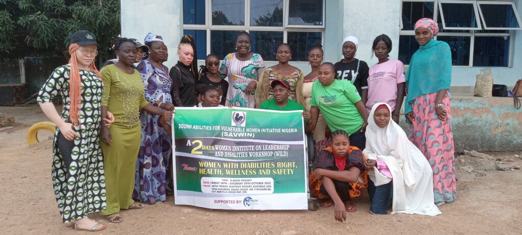 Nearly 20 women with disabilities, all participants of WILD-Nigeria, sit and stand together with WILD leader Miyausa holding a banner. The banner says "A 2 day WILD Workshop. Theme: Women with Disabilities Rights, Health, Wellness and Safety."