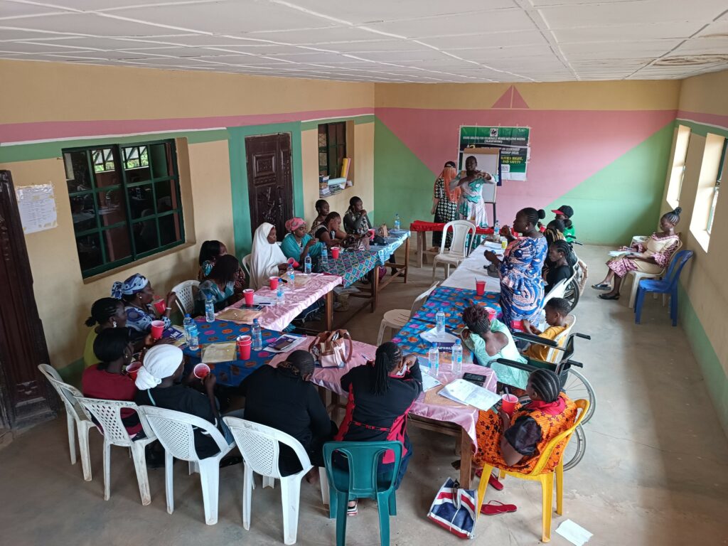 WILD-Nigeria leader Miyausa presents using sign language. 20 women with various disabilities sit together around long tables in the room.