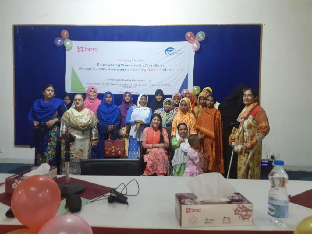 A group of women with various disabilities, all participants of WILD-Bangladesh, smile and pose for a group photo. Behind them is a banner with colorful balloons taped to the corner. The banner includes the words "Training Session: Empowering Women with Disabilities through building awareness on law, legislation, and advocacy" and includes logos of MIUSA and BRAC.