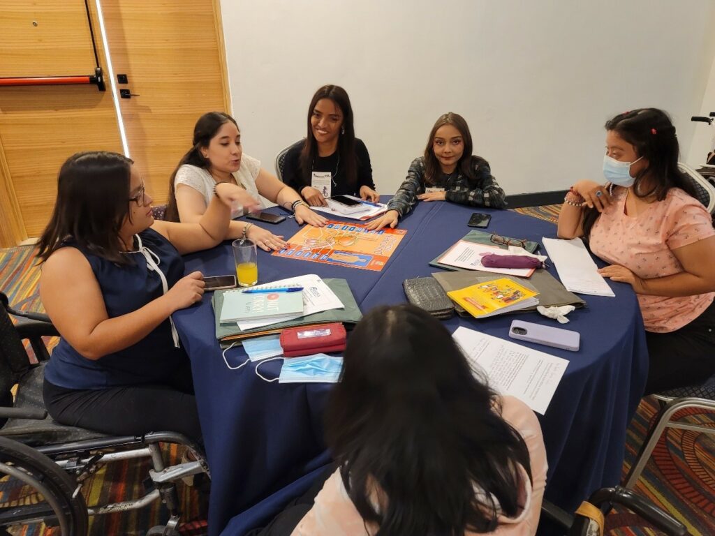 Six women participants of WILD-El Salvador, some wearing facemasks, sit around a round table with paper materials on the table. One participant sits in a manual wheelchair.
