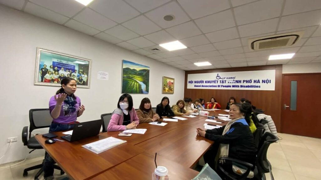 WILD-Vietnam leader Giang stands near a laptop computer while speaking to a group of women participants. They are sitting at a long table in a conference room.