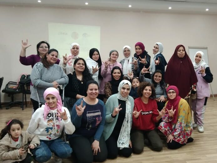Twenty women with various disabilities stand or kneel together facing the camera. Many are wearing headscarves, smiling, and holding up one hand gesturing the sign for "I love you."