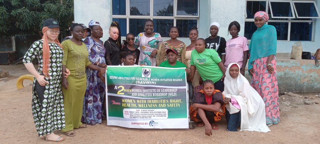 Twenty women with diverse disabilities sit and stand next to each other outside of a building. A few women are holding  a banner that has SAVWIN's logo and words stating "A 2 Day Women's Institute on Leadership and Disabilities Workshop (WILD)" and "Theme: Women with Disabilities Rights, Health, Wellness, and Safety".