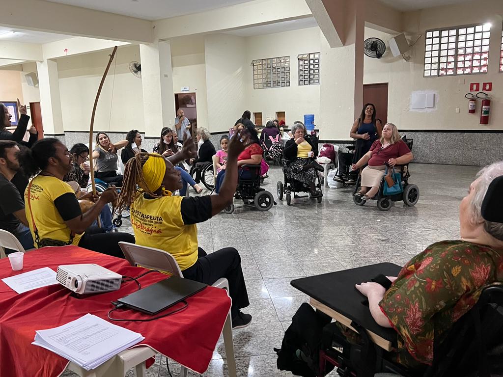 Women with various disabilities gather in a room, facing two guest presenters in yellow shirts. The two presenters have their arms raised and musical instruments on their laps.