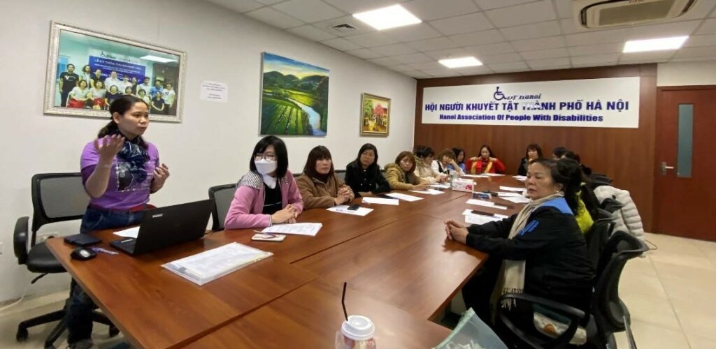 Several woman sit around a long conference table facing a presenter. The presenter (Giang) is standing in front of a laptop computer and speaking to the group.