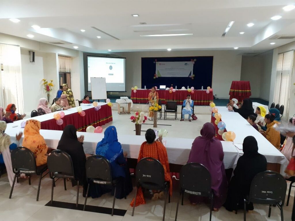 Twenty women with disabilities, some wearing headscarves, sit around a U-shaped conference table facing Sharmin. Sharmin presents with a microphone facing participants.