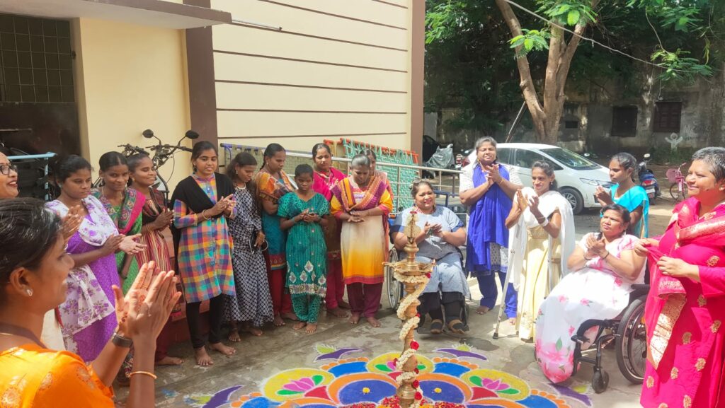 A group of 18 women in India with various disabilities gather in a semi-circle outdoors, with a large colorful art design on the ground in front of them. Many are smiling and some are clapping their hands together. 