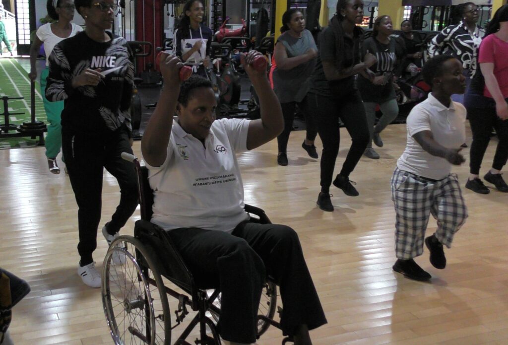 A person sitting in a manual wheelchair holds two weights over their head in a large gym facility. Behind them and beside them are several other women with disabilities also doing the aerobics activity, standing lifting one foot off of the ground.