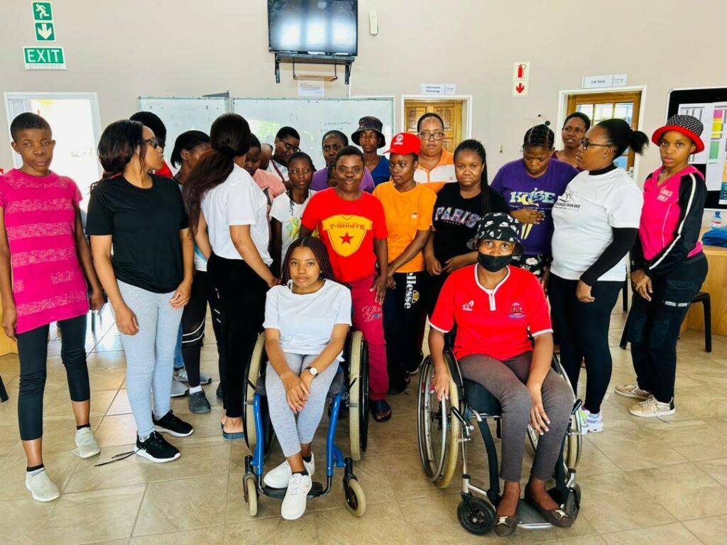 25 women with various disabilities, some using wheelchairs, sit and stand together in a classroom facing the camera.