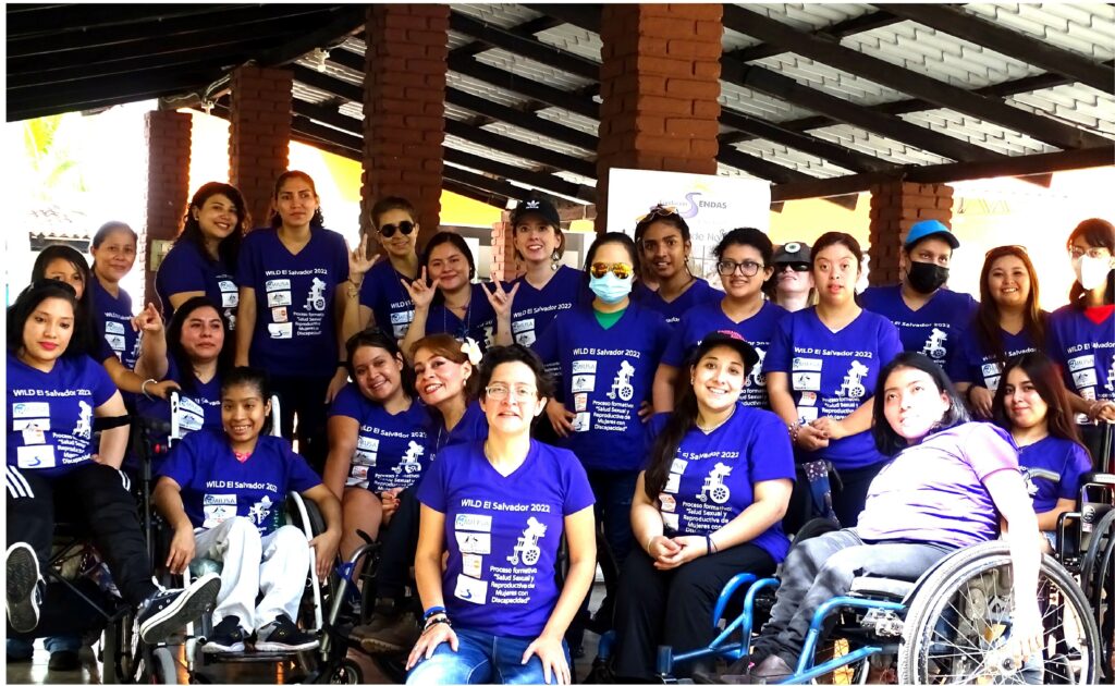 22 women with diverse disabilities, some using wheelchairs or other mobility aids, all wear bright purple t-shirts. They are smiling facing the camera while posing under an outdoor shelter.