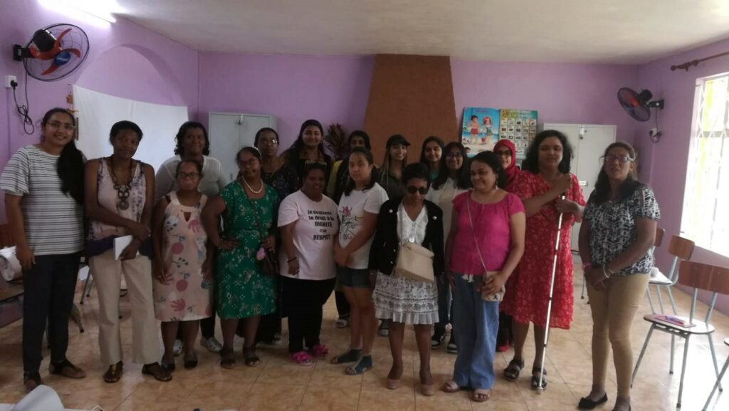 A group of women with various disabilities stand next to each other in a classroom smiling for a photo. The lead trainer, Aarthi, wears a bright red dress and holds her white cane.