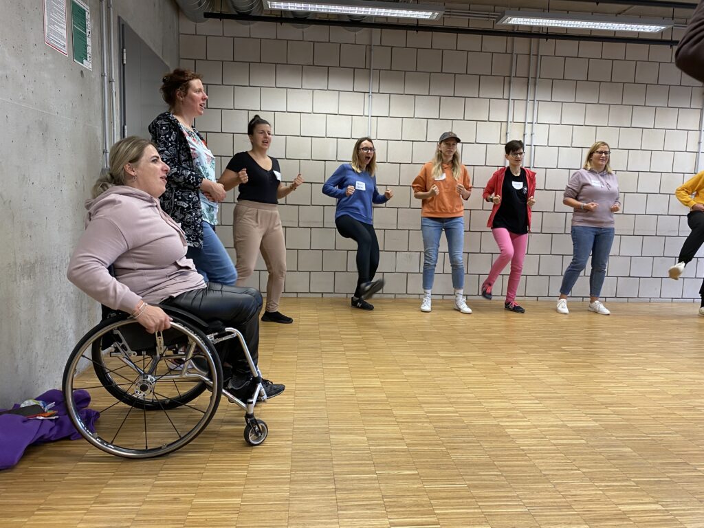 Several women, including one using a wheelchair, sit or stand in a large empty room during self-defense. Some are shouting while kicking one foot forward and holding two fists.
