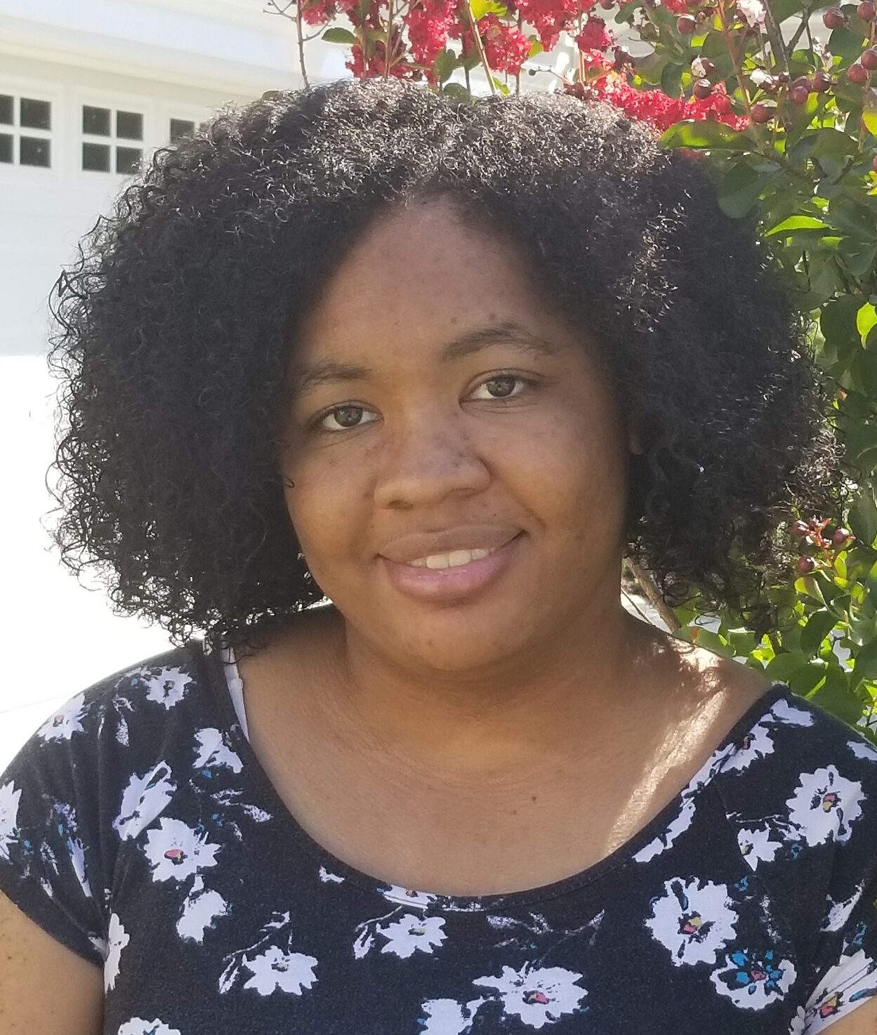 Zhane is a young black person with curly black hair wearing floral top and standing in front of flowers, smiling at camera.