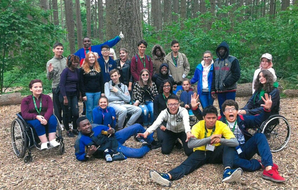 Group of high school students smiling while in an outdoor park. Behind them are tall trees and green plants. Some students are using manual wheelchairs.