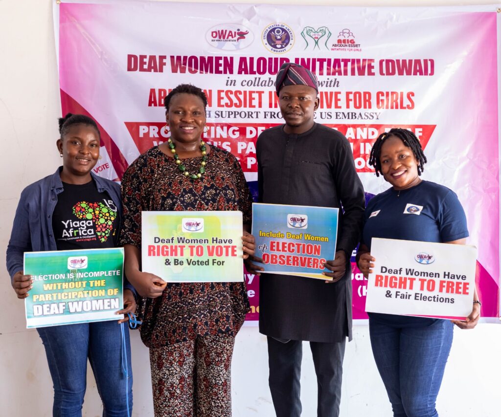 Four people stand side by side in front of a banner that reads "Deaf Women Aloud Initiative" and other obscured text. Each person holds a sign that says "Deaf Women Have Right to Vote" and variations