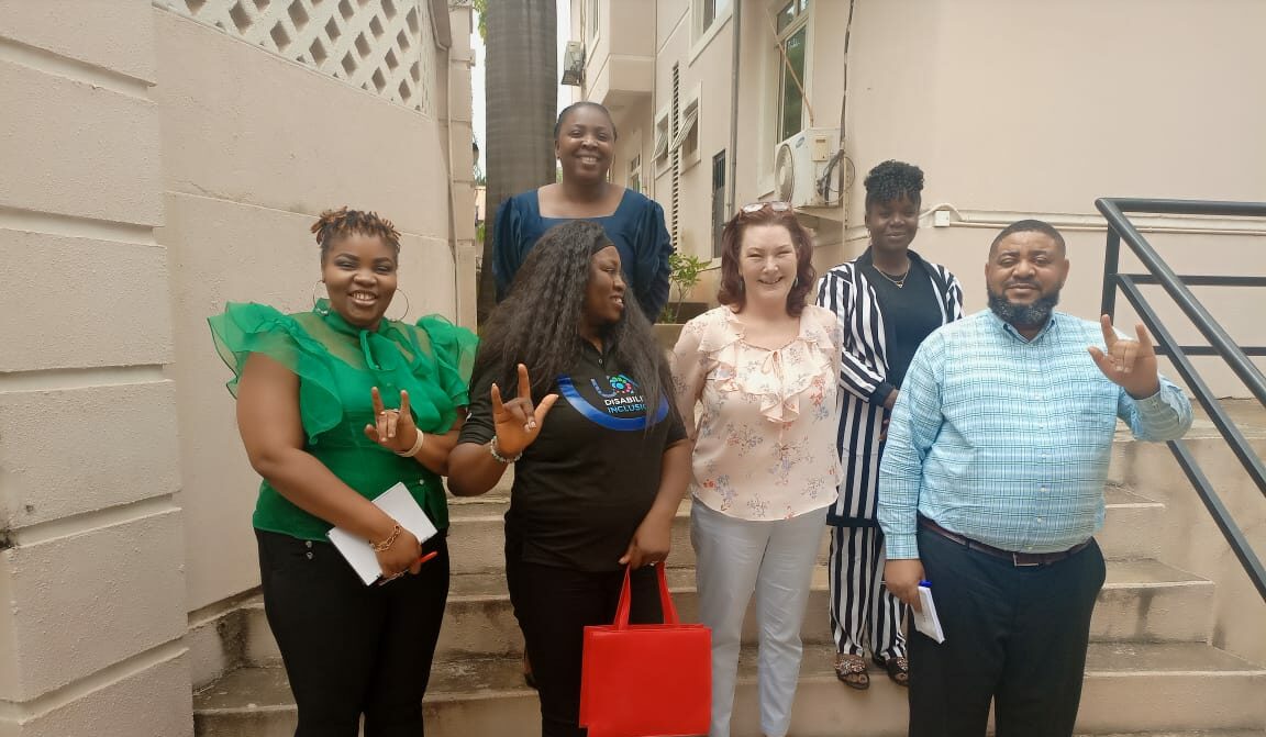 A group of six people stand on an outdoor staircase in two rows; Some people make the "I love you" hand shape in sign language. They wear business casual attire."