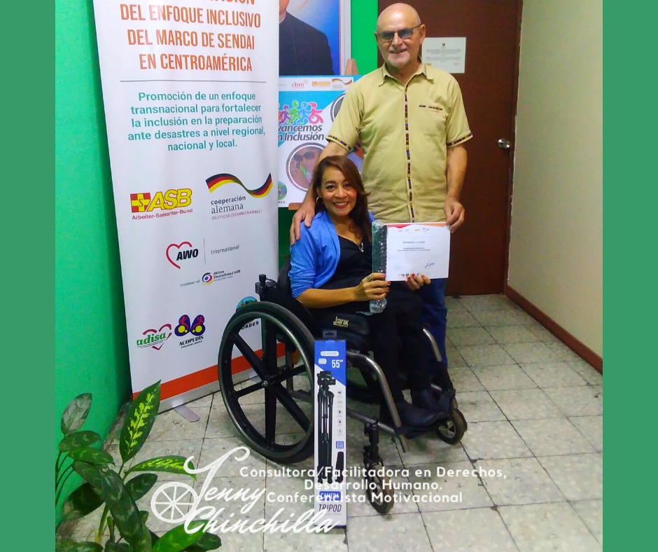 Jenny sits in her wheelchair holding a certificate of first place award for her documentary. In front of her is a camera tripod in a box. A man stands to Jenny's left with his hand on her shoulder. To Jenny's right is a sign displaying funders of the project and project title in Spanish.