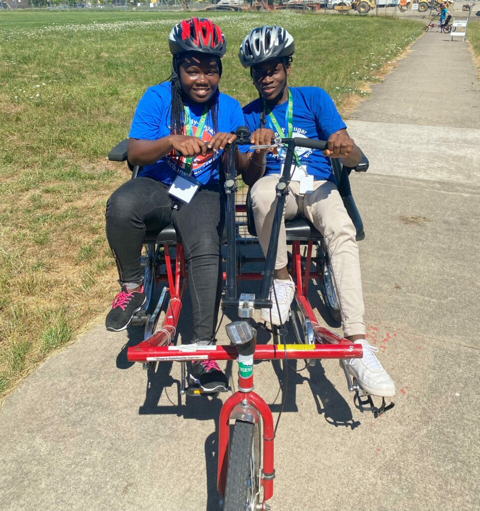Two students wearing blue YES programs shirts sit side by side on a tandem tricycle smiling.