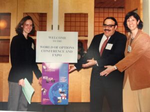 Vintage photo of David Levin in perhaps his 40s, wearing suit and tie and moustache, standing indoors pointing to a sign that says Welcome to a World of Options Conference