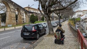 Man rising a powerwheel chair seen from behind on a car-lined cobblestone sidewalk. Stone arches are in the background.