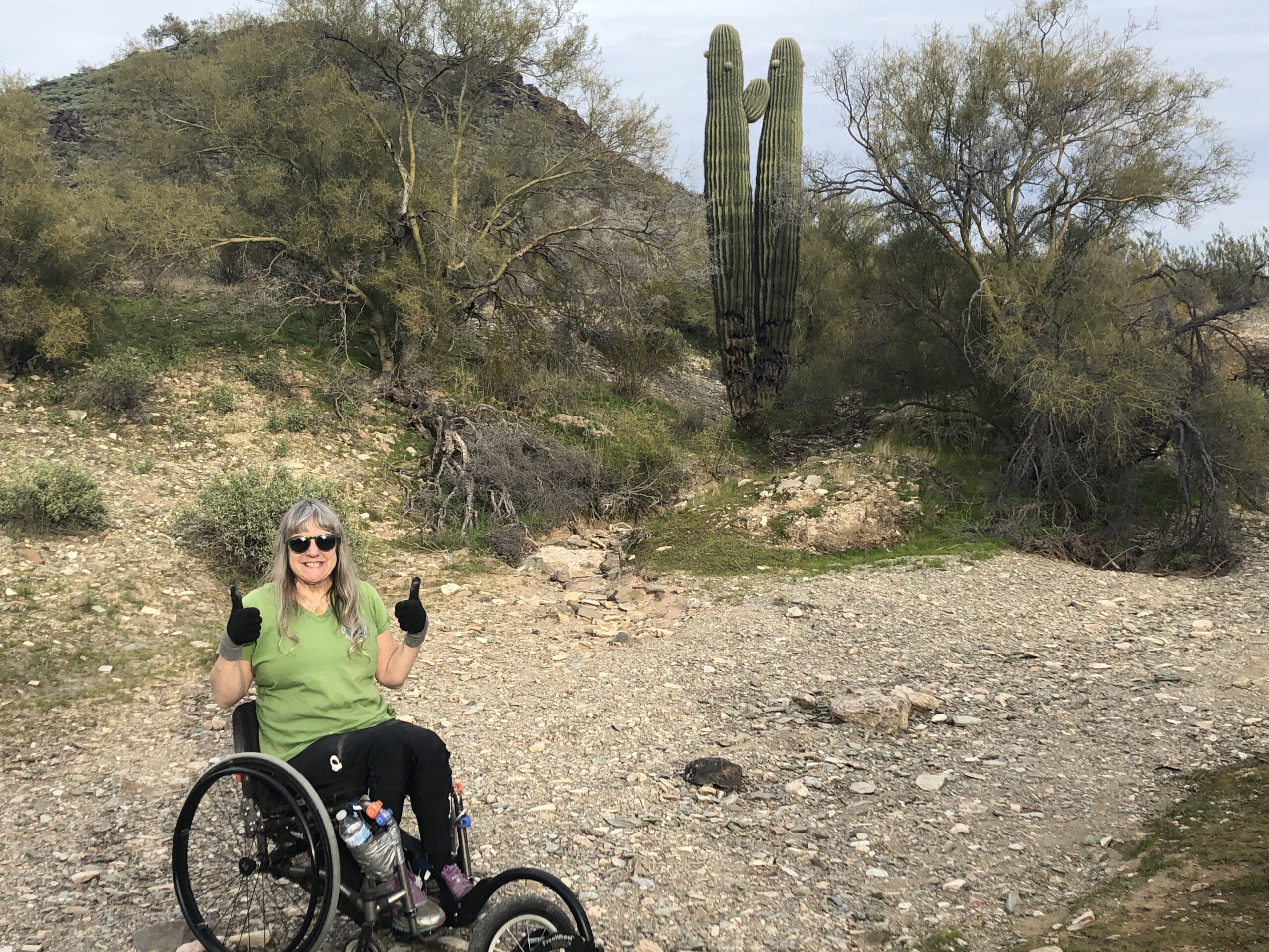 Susan gives thumbs up as she sits in her manual wheelchair with Freewheel front attachment, in a desert setting with cactus and hill. Susan wears sunglasses and gloves