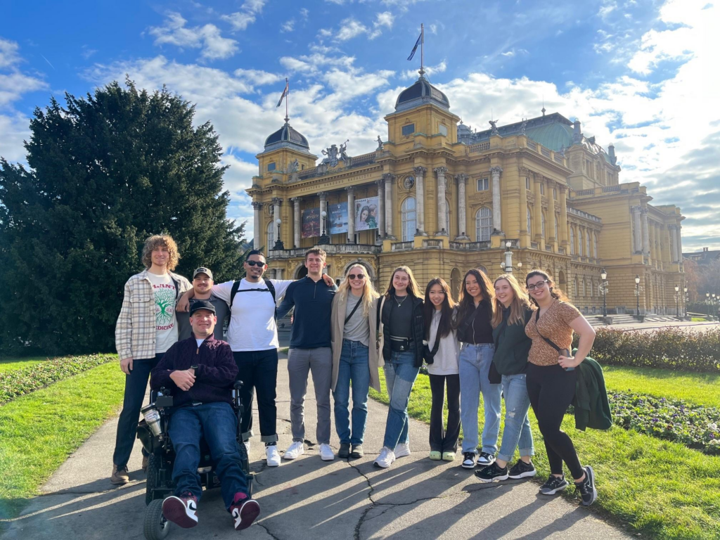 Group of college students standing with man riding power wheelchair in front of a building in Croatia.