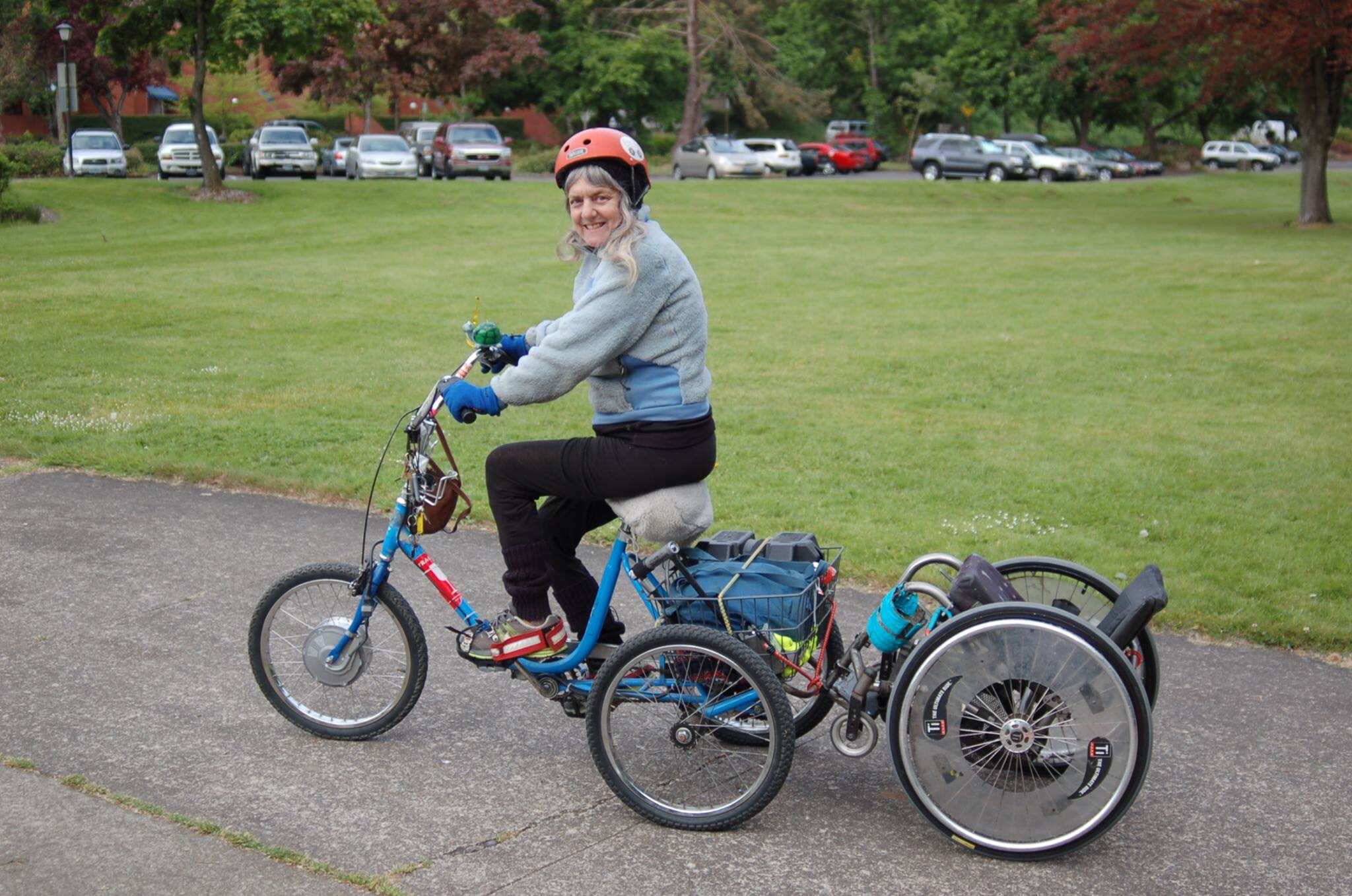 Susan rides an adult tricycle, with her manual wheelchair attached to the back of it. She wears a helmet and looks over her shoulder to smile at the camera.
