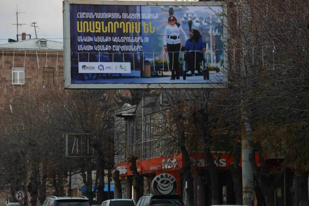 Billboard with Armenian text and an image of a blind woman walking with a white cane next to another women riding a power wheelchair. 