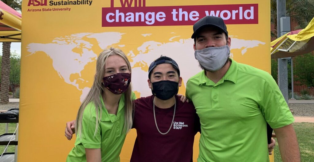Three people stand with arms around shoulders facing camera. They wear face masks. Kevin stands in center in ASU logo shirt. Behind them is a banner of a world map that says Change the World