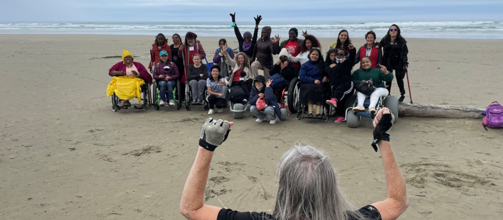 A group photo of WILD women on a beach with Susan Sygall raising cheering for them in the foreground.