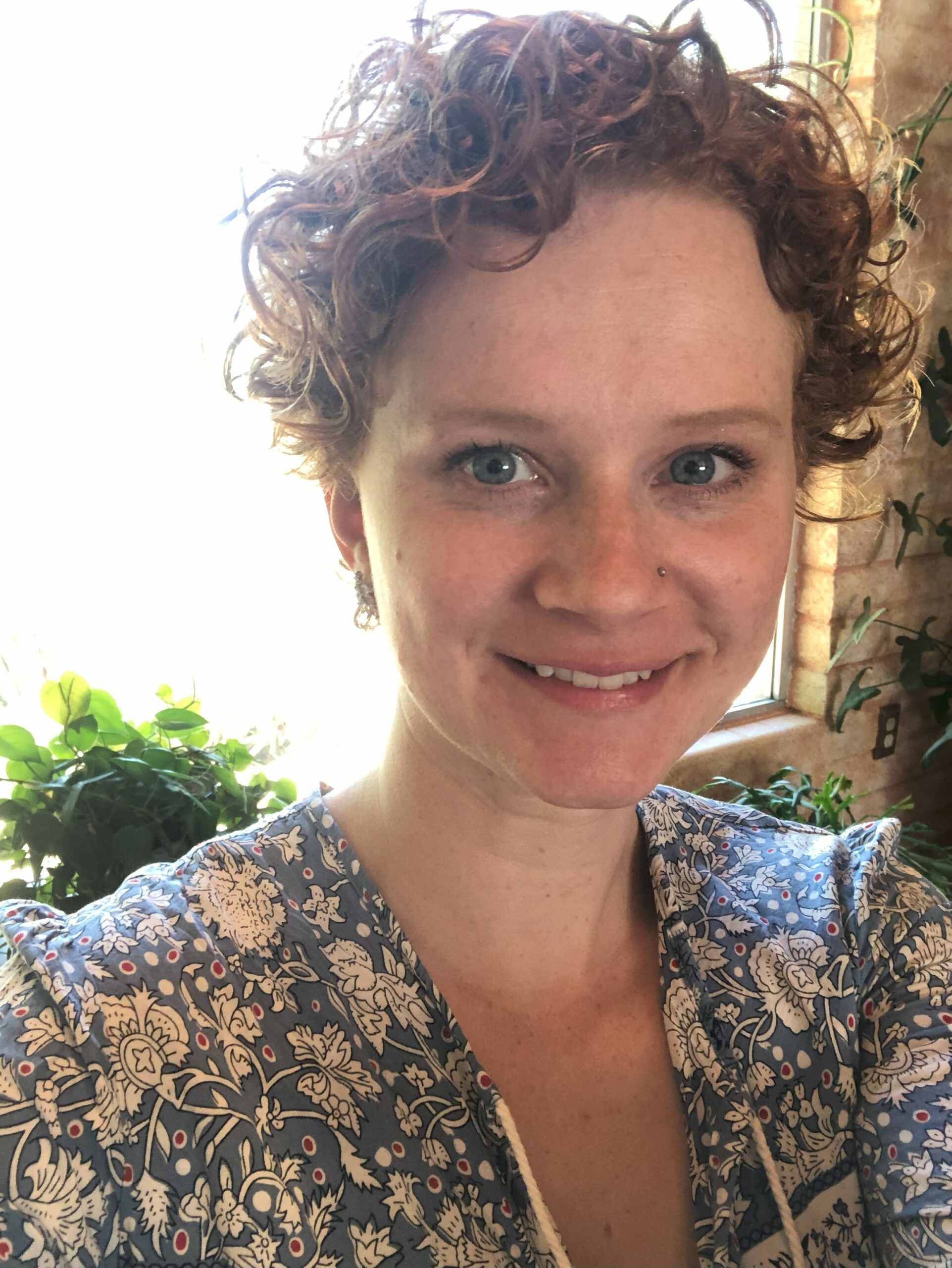 Fiona headshot smiling and looking at camera, young white woman with short hair and floral top, bright window in background
