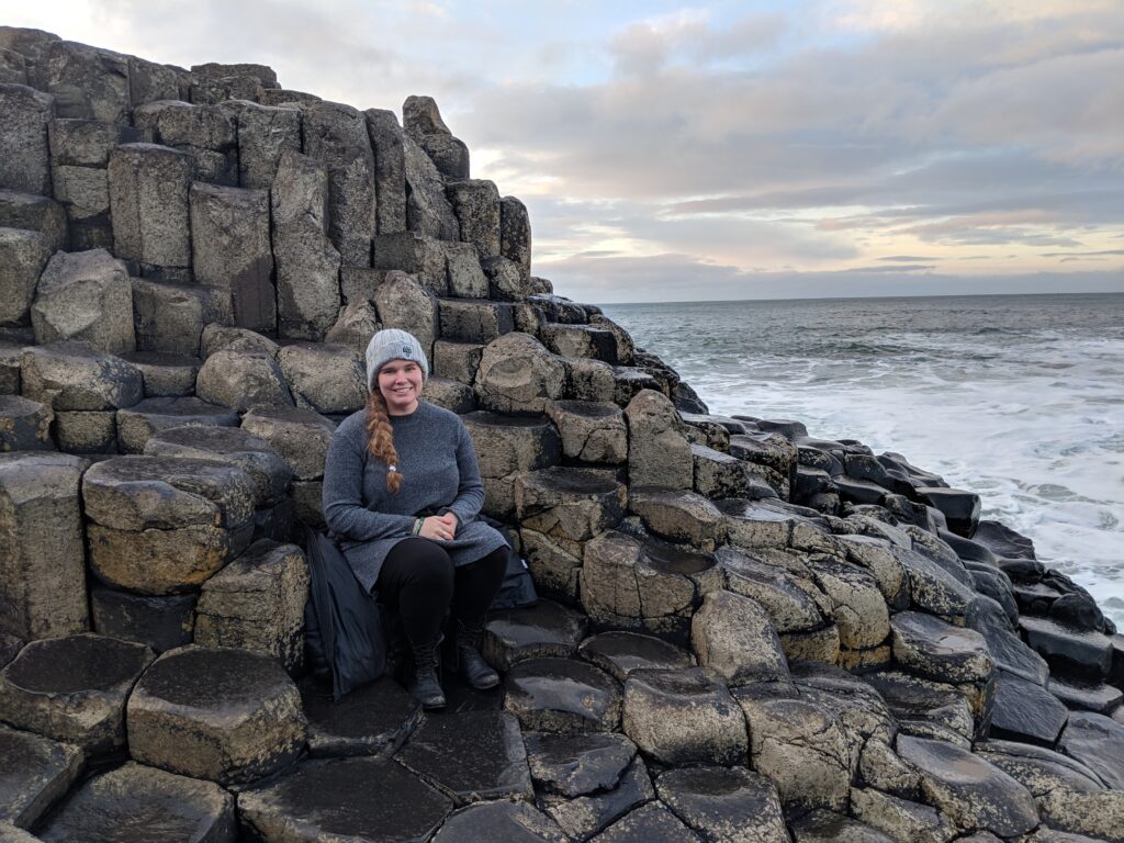 Lindsey wears sweater and knit hat, sits on a rocky shore next to the sea and cloudy sky