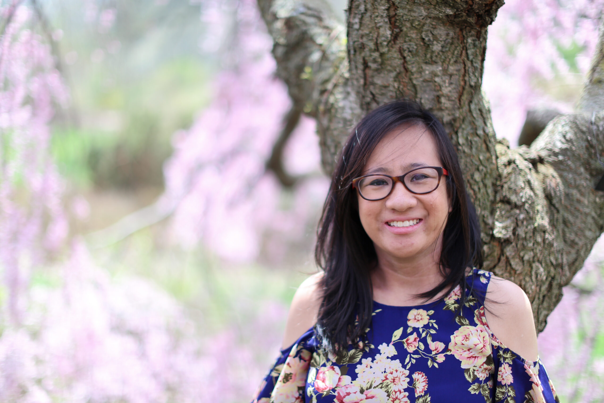 Geraldine is a young woman with Asian heritage. She stands in front of a blooming cherry blossom tree smiling at camera.
