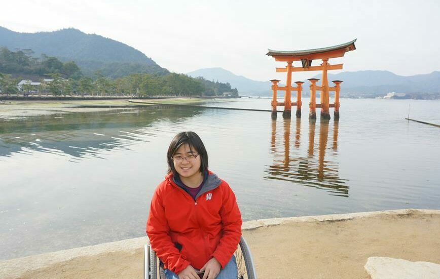 Alahna is a young Asian American woman seated in a wheelchair in front of a lake with a tall Japanese gate emerging from the water