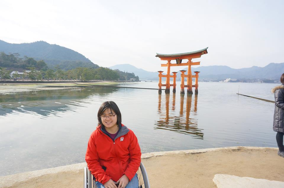 Alahna is a young woman with Asian heritage who sits in a wheelchair wearing red coat in front of a pool of a body of water out of which emerges a Japanese gate
