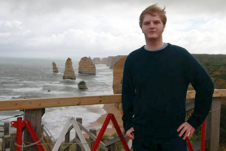 Kevin is a young white man who stands in front of a rail where behind him is a beach with large rocks emerging from the waves