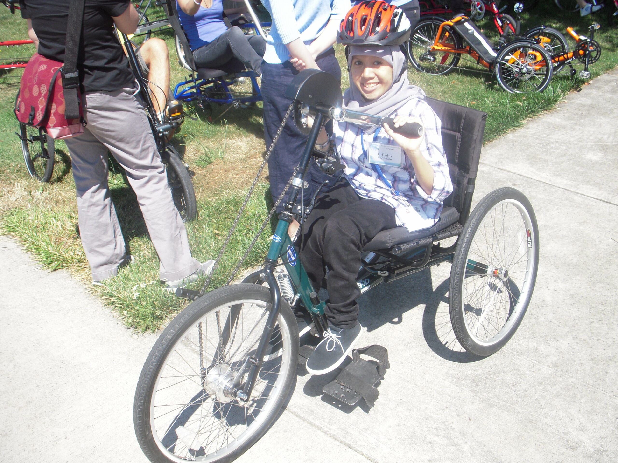 Lintang is a teen girl of southeast asian heritage. She wears a helmet over her headscarf and sits in a three-wheeled adaptive bicycle on a sidewalk with people behind her.