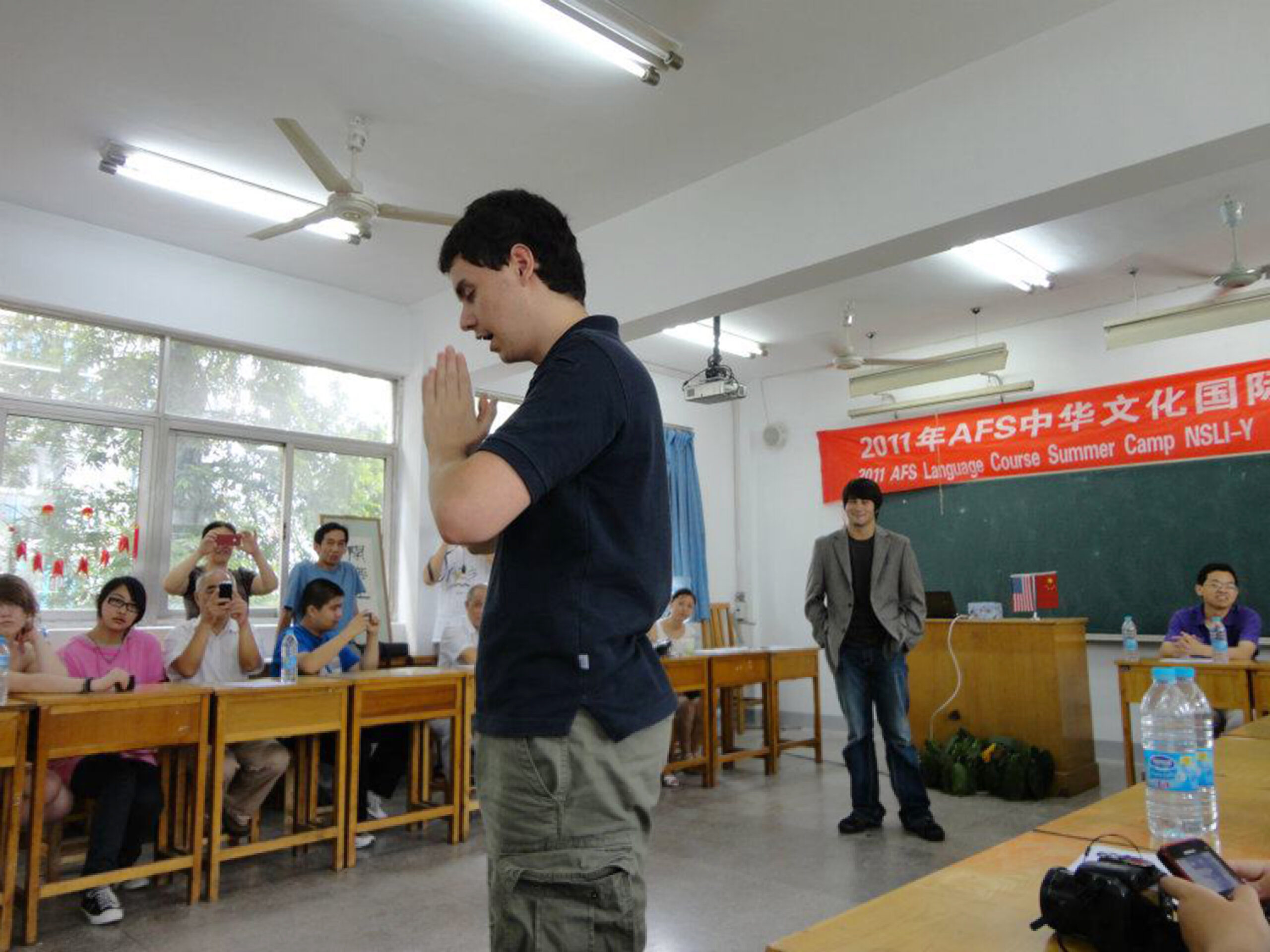 Nathan is a young white man with Asian heritage who stands in a classroom speaking to a group of Asian teachers and students. He bows his head and has his hands in a prayer shape. A banner has Chinese text and reads AFS NSLI-Y