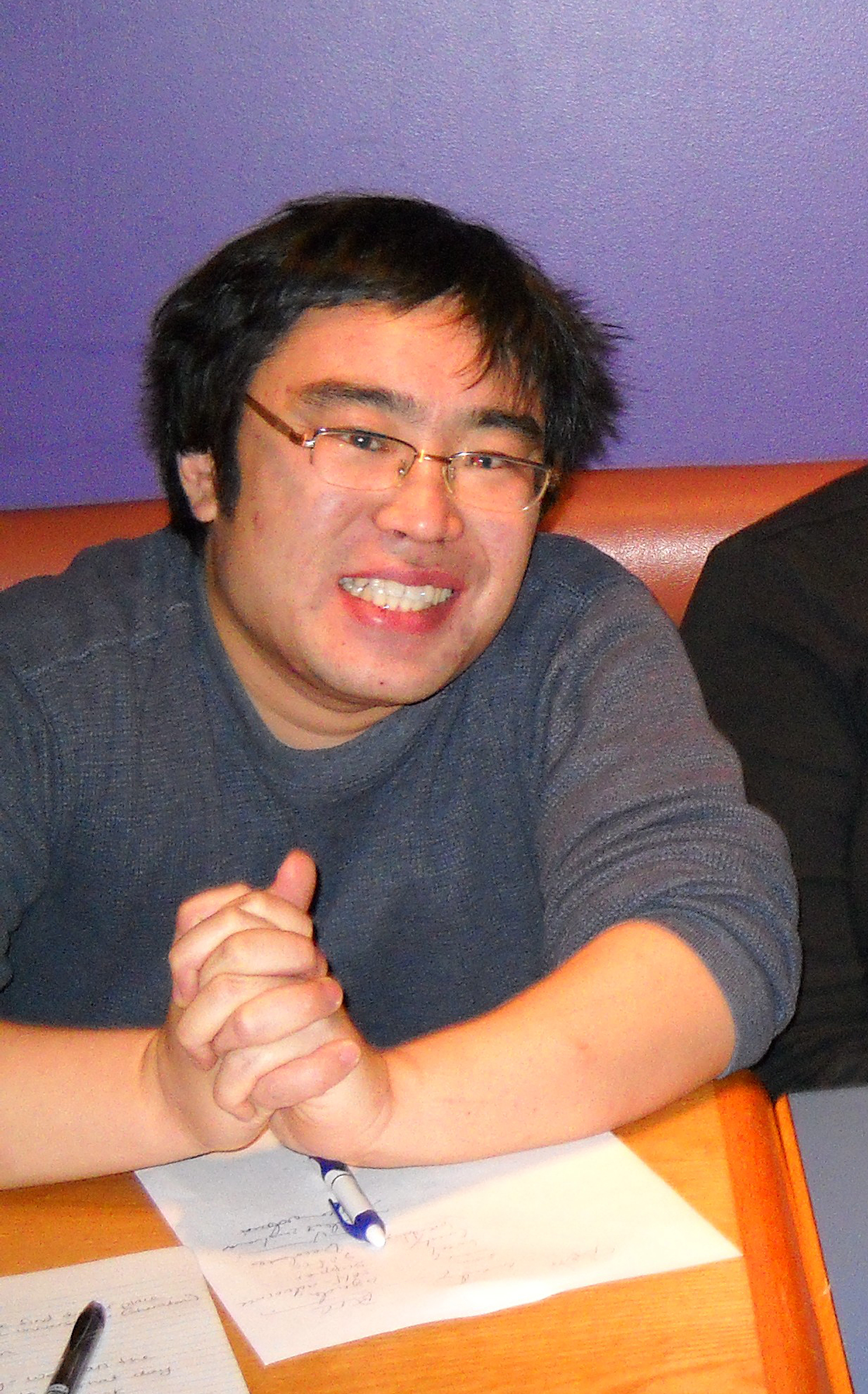 Yasushi is a young Japanese man who smiles at camera, leaning over a desk with hands clasped