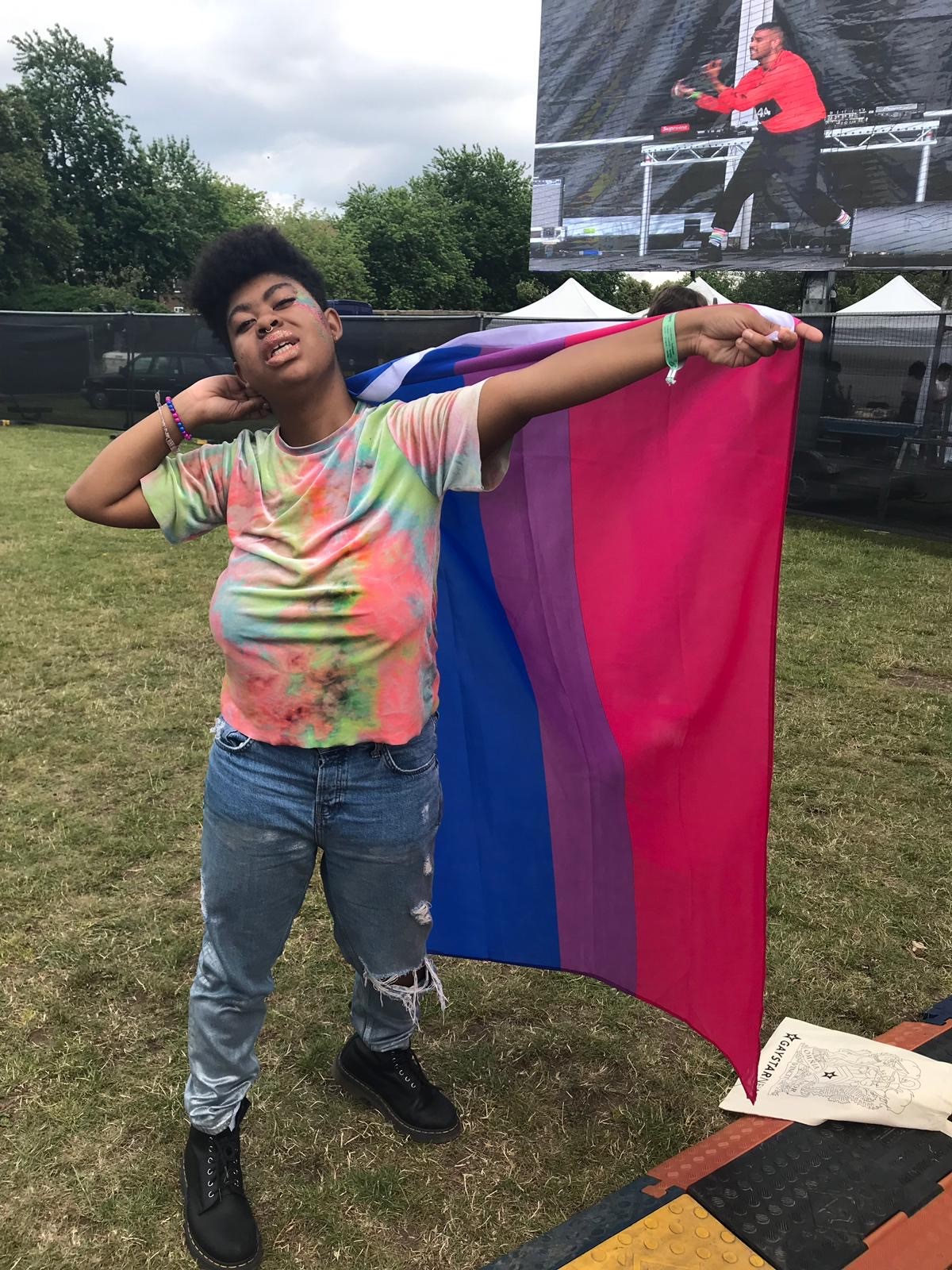 A young black non-binary person wearing tie dye t-shirt and jeans holds a flag with pink, purple, and blue stripes