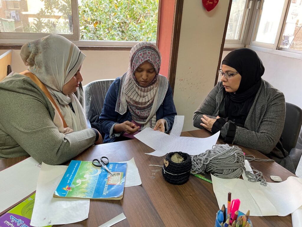 Three women wearing headscarves sitting around a table working on a project with scissors and paper.