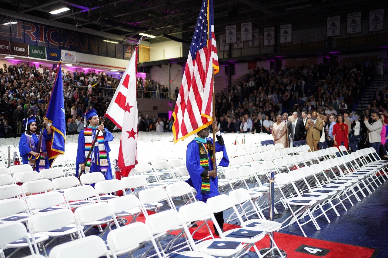 Julius wears graduation cap and gown and walks through rows of empty white chairs while carrying American flag. Other graduating students follow behind him.