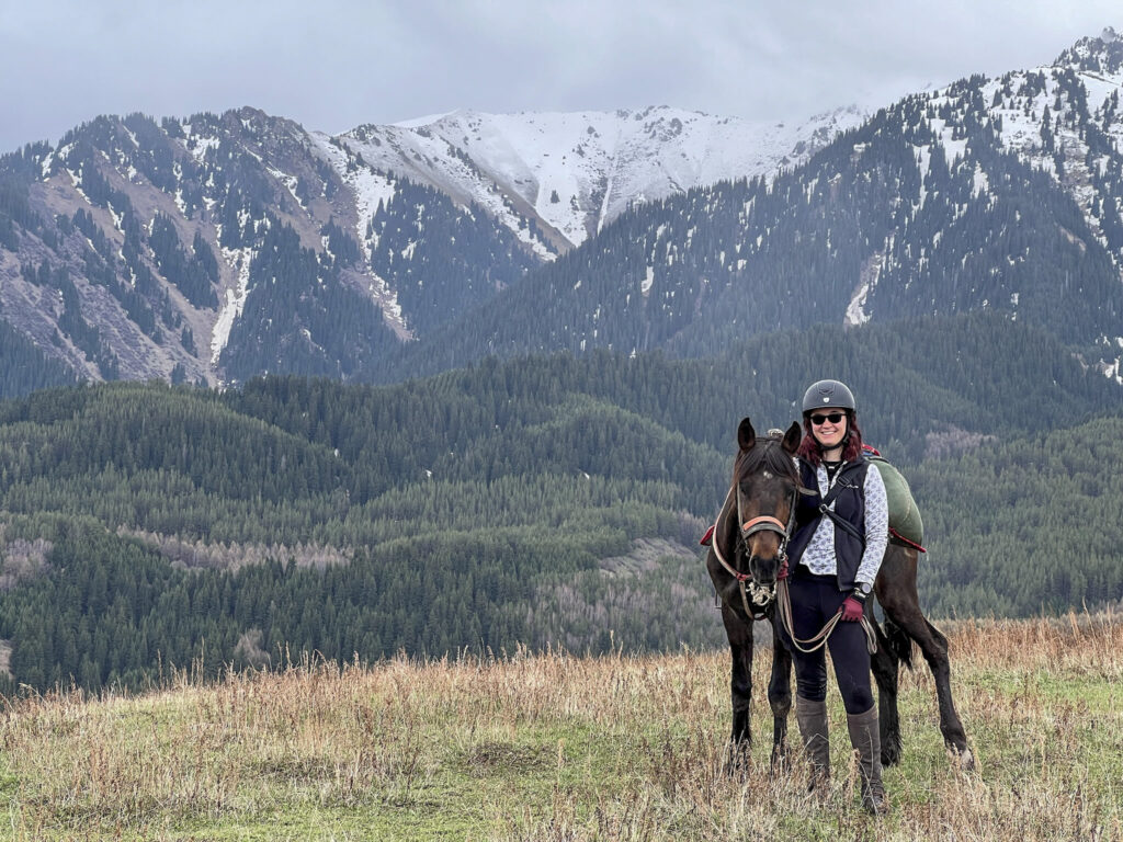 Picture of a woman standing next to a dark brown horse in a mountainous area wearing a helmet, sunglasses, a patterned long-sleeve shirt, a black vest, gloves, and riding boots.