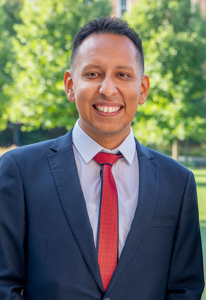 A professional portrait of Michael wearing a suit and tie, standing outdoors and smiling.