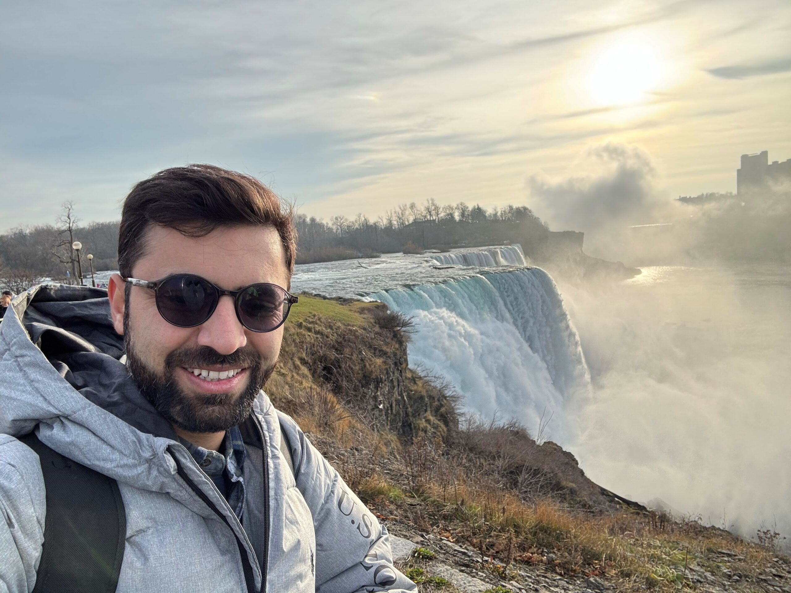 A young man wearing sunglasses and a goatee stands smiling at camera by a railing with Niagara Falls in background, golden in the sun and mist