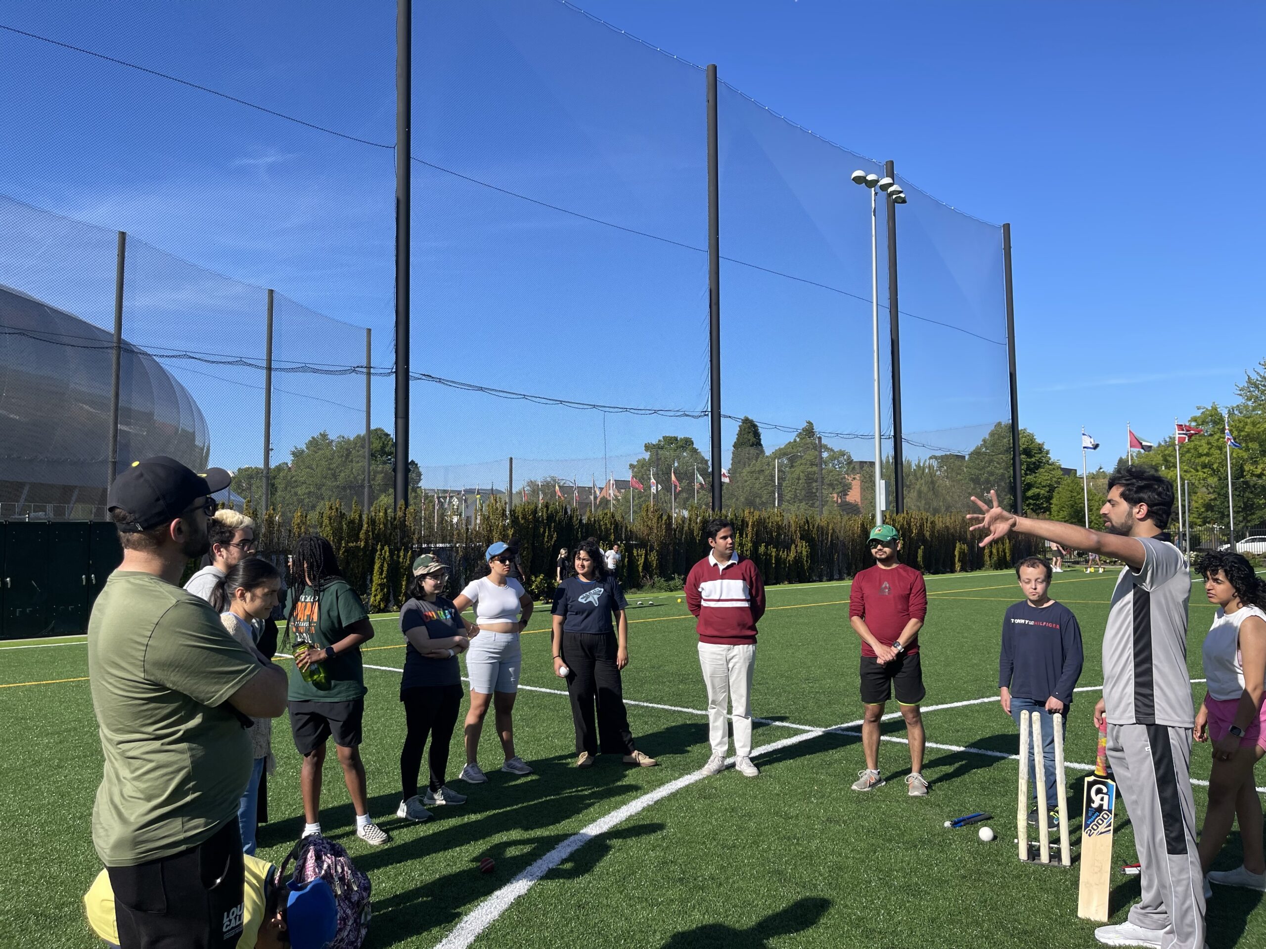 On an outdoor athletic green lined with tall fence, a small group of people stand in a circle while Sohaib (at far right) explains rules of cricket while holding cricket equipment