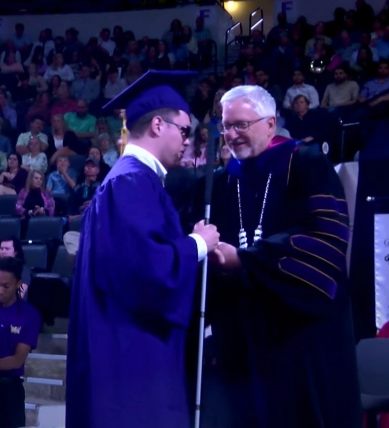 Vadim wears blue cap and gown and holds white cane in his hand as he shakes hands with an older man in black regalia.