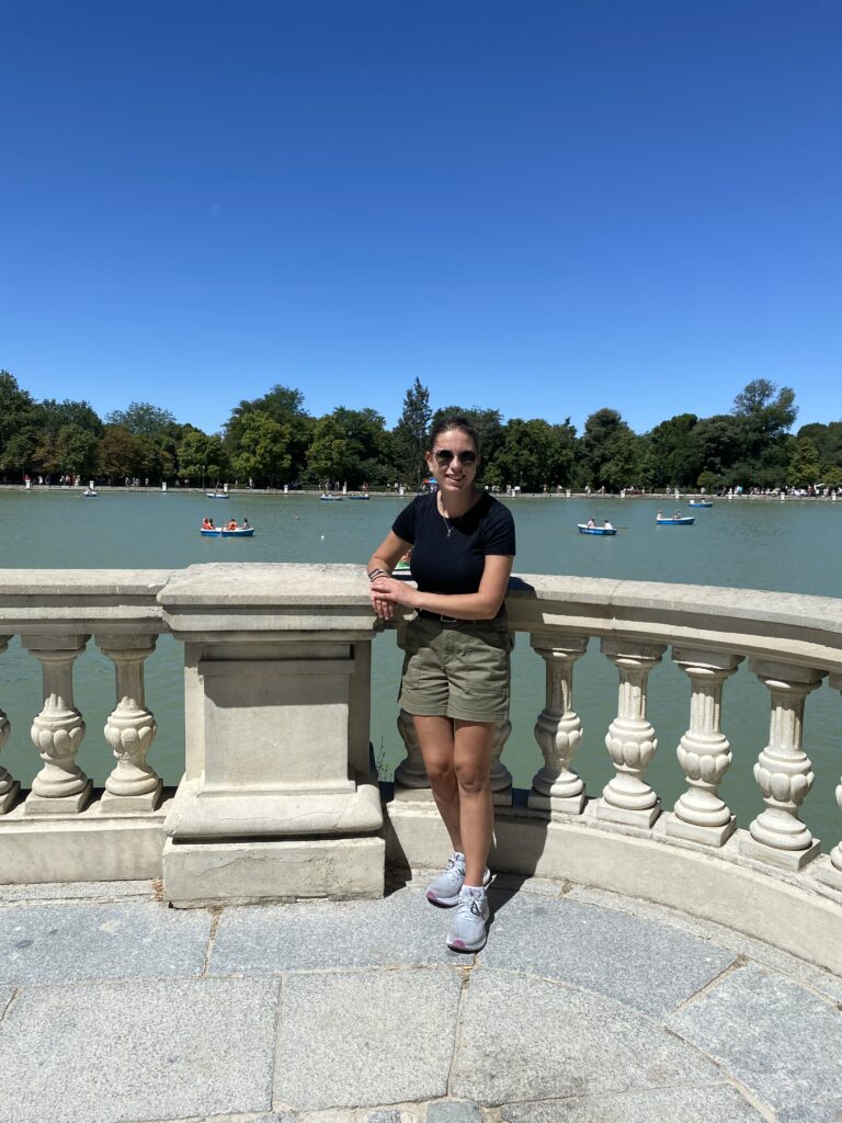a woman standing outdoors on a sunny day,leaning against a stone balustrade with a large body of water behind her. The woman is wearing a black t-shirt, olive green shorts, and gray sneakers. She has sunglasses on and is smiling at the camera. In the background, there are several small boats on the water with people in them, and a line of trees along the far shore. The sky is clear and blue.